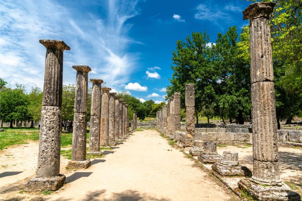 Ancient stone columns lined up under a bright blue sky with greenery surrounding the archaeological site at Olympia, Greece.