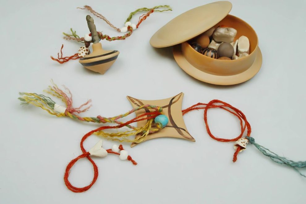 A selection of ancient Greek-inspired toys, including a spinning top, a string toy with colorful cords, and a lidded container holding small clay pieces and beads.