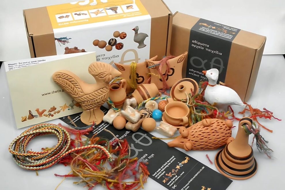 A collection of ancient Greek-inspired toy replicas, including clay figurines, vessels, and braided cords, displayed alongside informational packaging and colorful materials.