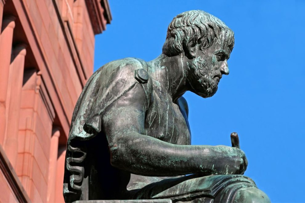 A bronze statue of Aristotle, the ancient Greek philosopher, shown in a seated position with a focused expression against a clear blue sky.