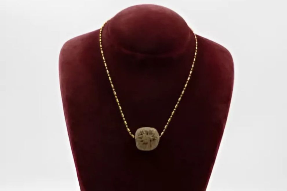 A handmade Ancient Greek-style necklace with a small carved pendant on a simple gold-tone chain, shown on a burgundy display