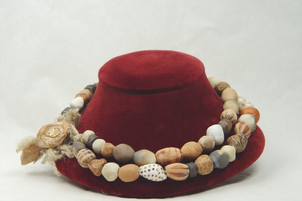 A handmade Ancient Greek-inspired necklace with various earthy-toned beads, displayed on a small red stand.