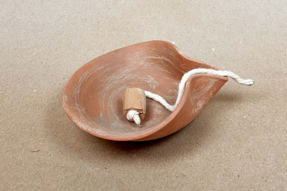 A hand-folded oil lamp from the Eastern Mediterranean, made of terracotta with a simple, curved design. A wick is threaded through a small clay holder, resting inside the lamp_old greek pottery