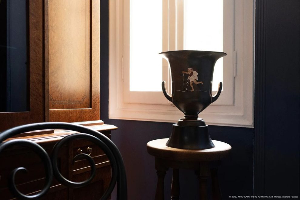 A South Italian Konakis Krater is placed on a wooden stool near a window, with soft natural light illuminating the scene. The dark pottery features a painted figure and has two handles. In the background, a wooden cabinet and part of a wrought iron chair are visible, adding to the classic interior setting.