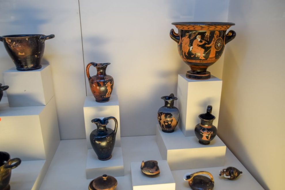 A display of ancient Greek pottery, including various decorated vases and vessels, arranged on white pedestals in a museum setting.