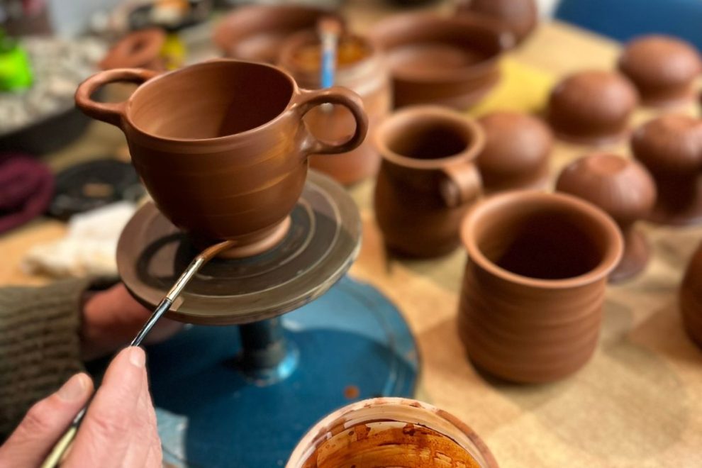 A potter’s wheel at Attic Black Studio, with a craftsman painting a clay cup. Various pottery pieces are visible in the background.